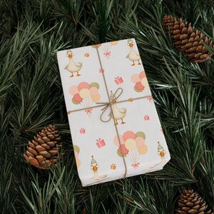 May include: A wrapped gift with a white background featuring a pattern of cartoon geese wearing party hats, balloons, and gift boxes. The gift is tied with twine and set against a backdrop of pine needles and pine cones.