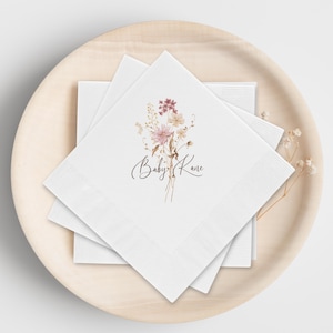 May include: White square napkins with a floral bouquet design in pink, beige, and brown tones. The text "Baby Kane" is written in a delicate script. The napkins are stacked on a wooden plate.