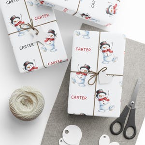 May include: White wrapping paper featuring a repeating pattern of watercolour snowmen wearing black top hats and red plaid scarves, interspersed with the name Carter printed in red or black text, tied with brown twine and a star-shaped tag.
