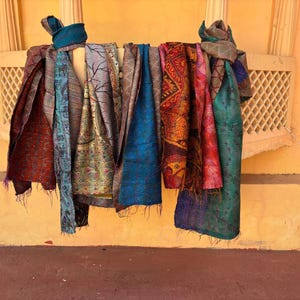 May include: A collection of colorful scarves hanging on a rod against a yellow wall. The scarves are made of various materials and feature intricate patterns in shades of red, blue, green, and gold. A blue turban is also visible.