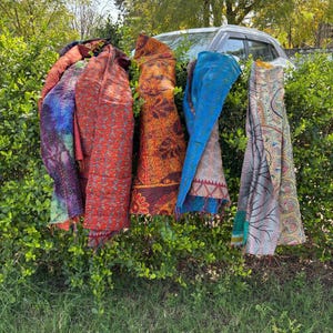 Puede incluir: Una colección de coloridas bufandas estampadas colgadas de un arbusto verde. Las bufandas muestran una variedad de colores, incluyendo rojo, naranja, azul y morado, con diseños intrincados. La imagen está tomada al aire libre, con un coche visible en el fondo.