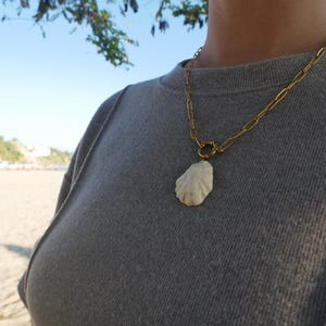 Op de afbeelding: Een goudkleurige ketting met een grote, gebroken witte schelpenhanger. De ketting wordt gedragen over een grijze trui. De achtergrond toont een strand en blauwe lucht.