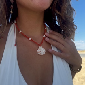 May include: A necklace featuring a red cord with a macrame knot pattern, accented by small white beads and a central pendant made from a flat, mottled white and tan seashell. The necklace includes a thin adjustable cord with a small bead at the end.