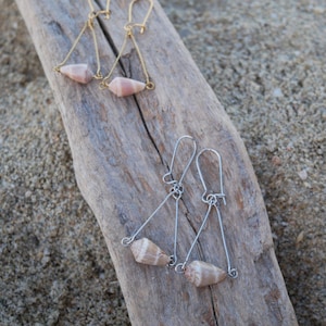 Può includere: Due paia di orecchini fatti a mano con conchiglie naturali. Un paio ha filo color oro e l'altro color argento. Gli orecchini sono di forma triangolare, con una conchiglia sospesa in fondo a ogni triangolo. Gli orecchini sono esposti su un pezzo di legno trasportato dal mare.