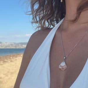 May include: A silver-toned chain necklace featuring a natural seashell pendant with white and reddish-brown coloration. The pendant is suspended from a simple metal bail, and the necklace is worn against a white garment with a deep neckline.