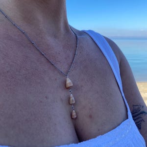 Op de afbeelding: Een zilveren ketting met drie kleine, lichtbruine, driehoekige hangers. De ketting wordt gedragen door een persoon met een wit topje en een blauwe lucht op de achtergrond.