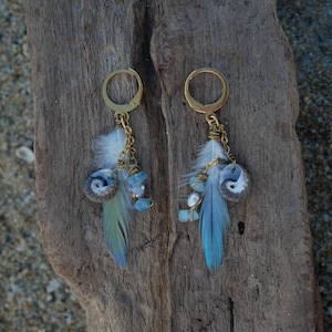 May include: A pair of gold-toned hoop earrings featuring dangling charms. Each earring includes a small spiral seashell, a white and blue-tipped feather, and several light blue and white beads suspended from a gold-toned metal chain link.