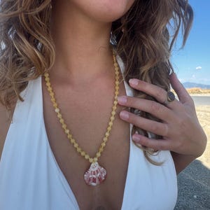 May include: A necklace featuring a strand of small, round, yellow beads and a single pendant shaped like a seashell. The seashell pendant has a white base with reddish-pink patterns and textures on its surface. The necklace is worn around the neck.