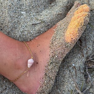 Könnte beinhalten: Ein goldfarbenes Fußkettchen mit einem rosa Edelsteinanhänger und einem kleinen, durchsichtigen Anhänger. Das Fußkettchen liegt auf einem mit Sand bedeckten Fuß, mit Strandhintergrund.