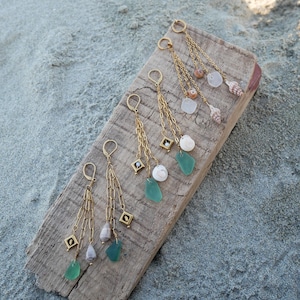 Könnte beinhalten: Mehrere Paare goldfarbener Ohrringe mit hängenden Ketten, Edelsteinen und Muscheln werden auf einem verwitterten Holzbrett präsentiert. Die Ohrringe haben grüne und weiße Edelsteine und kleine Muscheln.