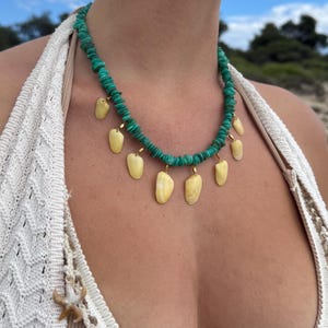 May include: A turquoise beaded necklace with shell pendants. The necklace is paired with matching earrings. The model is wearing a cream-colored crocheted top. The background shows a blue sky and trees.