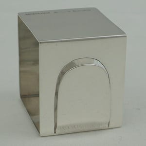 May include: A silver napkin holder in a cube shape with a curved cutout on one side. The polished metal reflects light, highlighting the clean lines and minimalist design. The napkin holder is likely made of stainless steel.