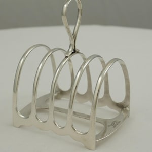 Puede incluir: Un tostador plateado con un asa curvada y cinco ranuras arqueadas. El tostador está hecho de metal brillante y tiene un diseño decorativo en la base. Un diseño clásico para servir tostadas.