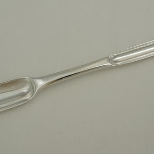 Könnte beinhalten: Ein langer, schlanker Marklöffel aus silberfarbenem Metall mit einer polierten, reflektierenden Oberfläche. Das Utensil hat an einem Ende eine schmale, längliche Laffe und am anderen Ende eine etwas breitere, abgerundete Laffe, verbunden durch einen glatten, konischen Griff.