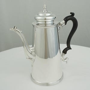 Puede incluir: Una cafetera plateada pulida con un asa negra. La cafetera tiene un pico largo y una tapa decorativa. El asa es curva y de madera oscura. La cafetera probablemente está hecha de acero inoxidable o un metal similar.