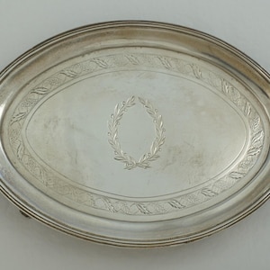 Puede incluir: Una bandeja de servicio ovalada de plata con acabado pulido y un borde decorativo con un patrón de hojas en relieve. El centro de la bandeja está grabado con un diseño detallado de corona de laurel, aportando un aspecto clásico a la pieza.