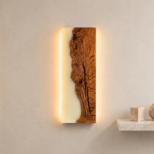 May include: A rectangular wall-mounted light fixture featuring a vertical slab of natural brown wood with a prominent grain pattern on one side and a smooth, translucent off-white resin panel on the other, emitting a warm glow from behind.