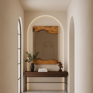 May include: A rectangular wall mirror with natural live-edge wood trim at the top and bottom, positioned above a dark brown wooden console table with thin black metal legs. A black vase with green foliage and a small abstract sculpture sit on the table.
