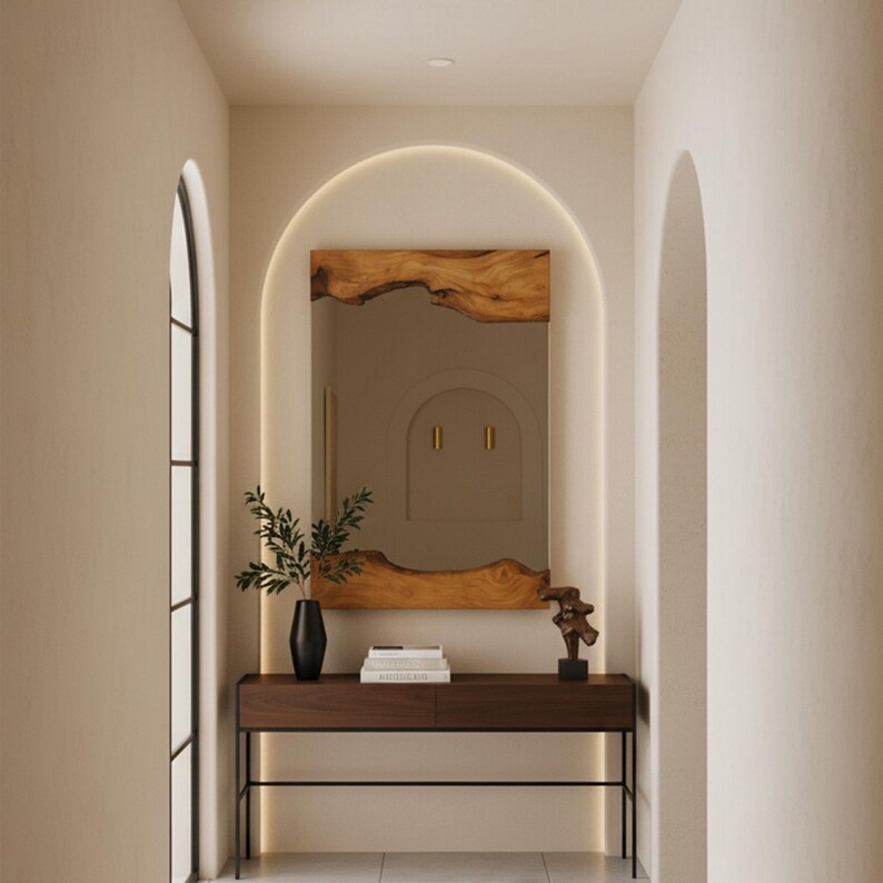May include: A rectangular wall mirror with natural live-edge wood trim at the top and bottom, positioned above a dark brown wooden console table with thin black metal legs. A black vase with green foliage and a small abstract sculpture sit on the table.
