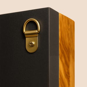 May include: A rectangular object featuring a matte black side panel and a natural wood grain side panel. A gold-toned metal D-ring hanger is securely attached to the black surface with a single matching gold-toned screw for wall mounting purposes.