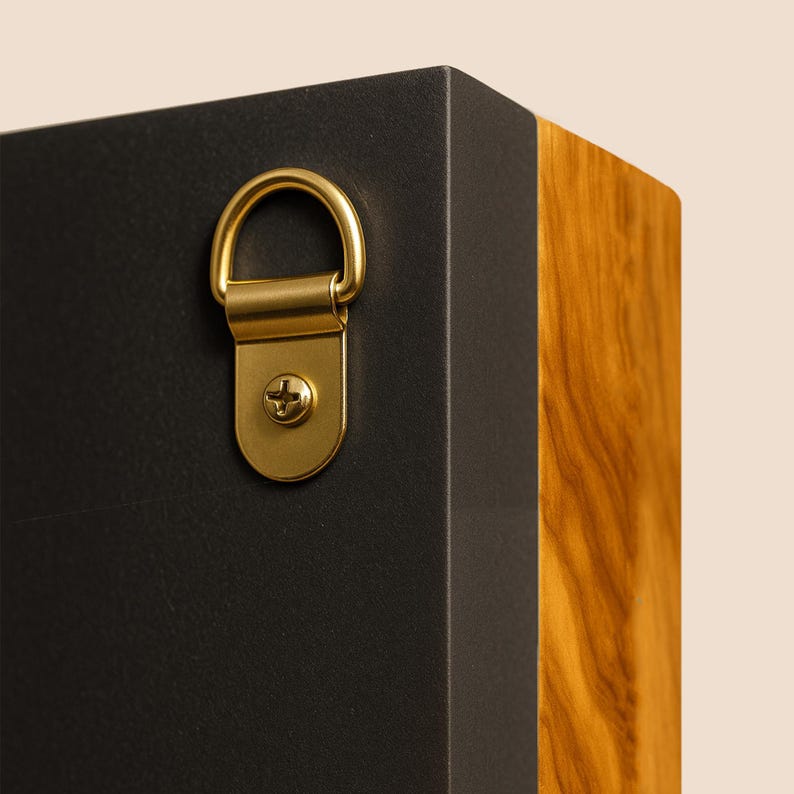 May include: A rectangular object featuring a matte black side panel and a natural wood grain side panel. A gold-toned metal D-ring hanger is securely attached to the black surface with a single matching gold-toned screw for wall mounting purposes.