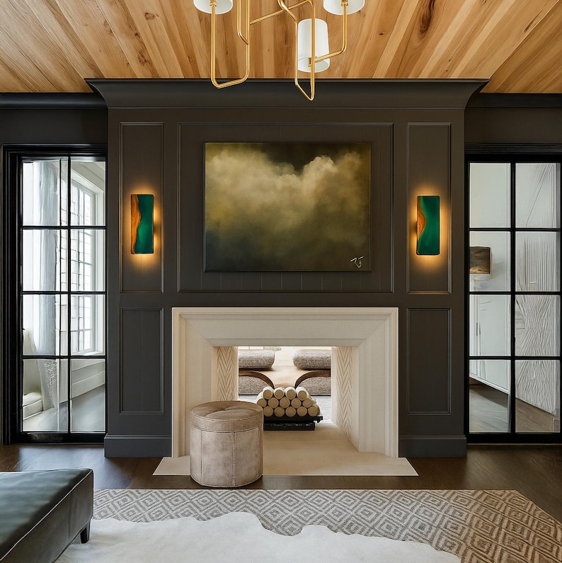 May include: A modern living room with a fireplace, artwork, and sconces. The fireplace is framed by dark gray paneling, with a painting above the mantel. Two green and gold sconces flank the artwork. A beige ottoman sits in front of the fireplace.