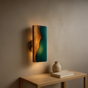 May include: A rectangular wall sconce with a wooden and teal resin design. The wood grain is visible on the left side, and the teal resin glows when lit. The sconce is mounted on a brass-colored circular base. A small table with a vase and book is below.