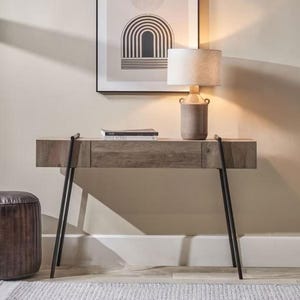 Puede incluir: Una consola de madera con acabado gris y patas de metal negro. Una lámpara beige con pantalla blanca está sobre la mesa, junto a una pila de libros. Una obra de arte abstracta enmarcada cuelga de la pared encima de la mesa.