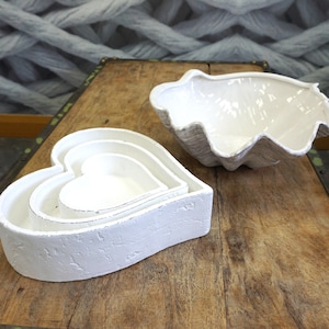 May include: White ceramic decorative bowls. One set is heart-shaped, with three nested bowls. The other is a large clam shell shape. The bowls have a textured, distressed finish, and are displayed on a wooden surface.