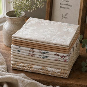 May include: A stack of folded cloth napkins in various neutral colours and floral patterns. The napkins are beige, tan, and brown. A framed sign behind the napkins reads "Create Something Beautiful". A vase of white flowers is to the left.