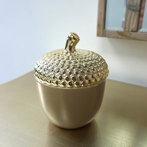 Pode incluir: Um recipiente em forma de bolota de cerâmica dourada com tampa texturizada. A tampa apresenta um padrão detalhado e borbulhante e um pequeno caule. O recipiente está sobre uma superfície dourada, adequado para decoração ou armazenamento.