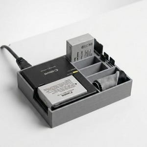 Könnte beinhalten: Grauer, 3D-gedruckter Kamera-Akku-Organizer mit einem Canon-Ladegerät und Akkus. Der Organizer hat mehrere Fächer für Akkus, ein Reinigungstuch und Speicherkarten. Ein schwarzes Netzkabel ist am Ladegerät befestigt.