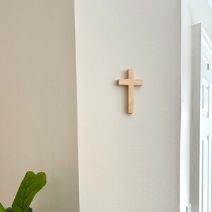 May include: A simple wooden cross mounted on a white wall, with a green leafy plant in the foreground.