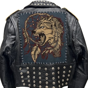 Op de afbeelding: Zwart leren jack met zilveren studs en een grote geborduurde patch op de rug. De patch toont een wolfskop met een droomvanger en veren.