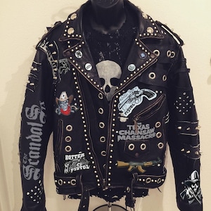 Peut inclure: Veste en cuir noir ornée de clous argentés, de pointes et d'écusson. Les écussons incluent une tête de mort avec un chapeau de cowboy, le texte "The Texas Chainsaw Massacre" et "Hotter than a $2 Pistol". La manche gauche affiche "The Scandal Fix".