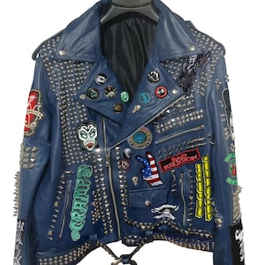 Puede incluir: Chaqueta de cuero azul con tachuelas plateadas y varios parches. La chaqueta tiene un diseño clásico con cuello de solapa, cierre de cremallera y múltiples bolsillos. Los parches incluyen una calavera, la bandera estadounidense y la inscripción "Bad Religion".