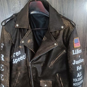 Könnte beinhalten: Eine braune Leder-Motorradjacke mit abgenutzten Details. Die Jacke hat einen Reißverschluss und einen Gürtel in der Taille. Der linke Ärmel hat die Worte "FREEDOM" und "free speech" aufgedruckt. Der rechte Ärmel hat die Worte "Liberty + Justice for All", "Tolerance", "Acceptance", und "Respect" aufgedruckt. Die Jacke hat außerdem einen kleinen amerikanischen Flaggen-Patch und einen Liberty Bell-Patch.