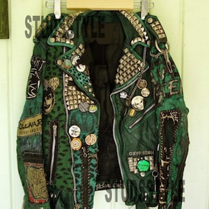 Op de afbeelding: Groene leren jas met zilveren studs, patches en spelden. De jas heeft een zwarte voering en ritsen. Patches met tekst als "OXYMORON" en "BEING THE".
