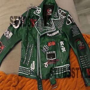 Può includere: Giacca in pelle verde scuro con borchie argentate, cerniere e toppe. Le toppe includono i testi "AC/DC", "The Art of War" e "Turisos". La giacca ha una cintura e un dettaglio a catena. Classico stile biker.