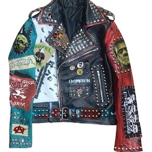 Peut inclure: Veste en cuir noire avec des panneaux rouges, blancs et turquoise. La veste est ornée de clous en métal, d'écussons et du mot "OXYMORON". Les écussons incluent un crâne, Frankenstein et les mots "EXTREME NOISE TERROR".
