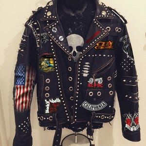 May include: Black leather jacket adorned with metal studs, spikes, and patches. Patches include the American flag, band logos like AC/DC, Ozzy Osbourne, and Guns N' Roses. The jacket has a zipper closure and belt details.