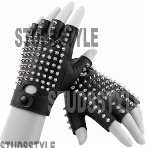 Puede incluir: Guantes negros sin dedos con tachuelas cónicas de metal plateado. Los guantes tienen un cierre de botón y un recorte cerca de la muñeca. Los guantes se muestran en manos de maniquí blancas.