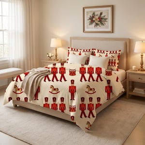 May include: A cream-colored duvet cover and matching pillow shams featuring a repeating pattern of red toy soldiers, rocking horses, and drums. A beige throw blanket is draped over the foot of the bed, which is centered between two bedside tables.