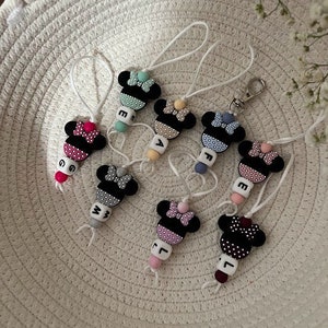 Może przedstawiać: Siedem kolorowych breloków silikonowych z czarno-białym wzorem Minnie Mouse. Każdy brelok ma koralik z literą z literami G, E, A, F, W, L i J.