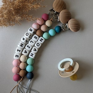 May include: A multi-coloured silicone baby teether with the names NOAH, ELIZA, and SOphia spelled out in white beads. The teether has two wooden clips and a matching pacifier.
