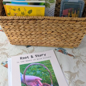 Puede incluir: Una cesta tejida llena de libros y un recipiente de plástico transparente con crayones. Un libro titulado "Root & Story In the Heart of Wild Violets" está en primer plano. La cesta y el libro están sobre una superficie con estampado floral.