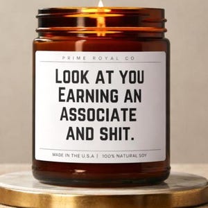 May include: A lit candle in an amber glass jar with a white label featuring the black text: PRIME ROYAL CO, LOOK AT YOU EARNING AN ASSOCIATE AND SHIT, MADE IN THE U.S.A | 100% NATURAL SOY. The candle sits on a round gold-rimmed marble coaster.