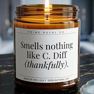 May include: A lit candle in an amber glass jar with a white label featuring the text Prime Royal Co. Smells nothing like C. Diff (thankfully). Made in the U.S.A. 100% natural soy. The candle has a warm glow and is filled with golden wax.