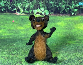 Pode incluir: Um pequeno urso de feltro marrom e preto com uma expressão feliz, em pé em uma superfície de grama verde. O urso tem um nariz marrom e olhos pretos.