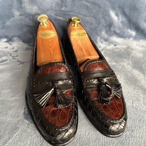 May include: A pair of black leather tassel loafers with brown crocodile-embossed leather accents. The loafers have a woven pattern and are on wooden shoe trees.